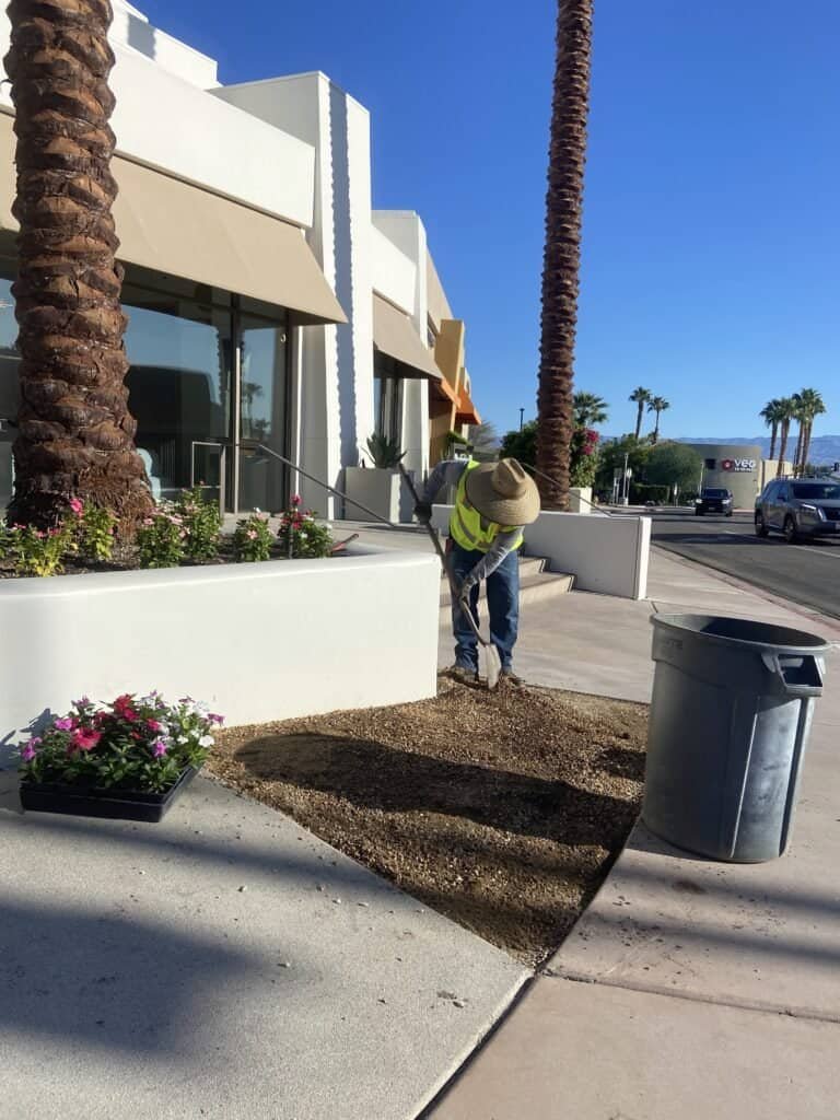Landscaping worker preparing soil for planting flowers near commercial building.